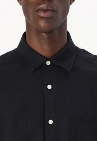 Sort tekstureret skjorte med en knaplukning på kraven, der har hvide knapper og en forlommer. Materialet virker let og åndbart.