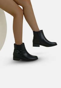 Botas de cuero negro hasta el tobillo con un acabado suave, cremallera lateral y hebilla decorativa, con punta redondeada y tacón bajo tipo bloque.