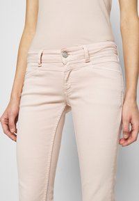 Jean rose clair à coupe slim, avec cinq poches et une fermeture à bouton argenté. Tissu lisse avec des détails de couture subtils.