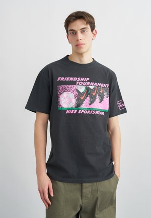 Fiatal férfi fekete Nike pólóban, melyen rózsaszín grafika látható focicipőkről, amik éppen rugdossák a labdát, valamint a "Barátság Torna Nike Sportswear" felirat.