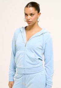 Ljusblå zip-up hoodie i mjukt tyg. Har en huva, framfickor, guldfärgade dragkedjeaccent och ribbade manschetter vid ärmarna och nederkant.