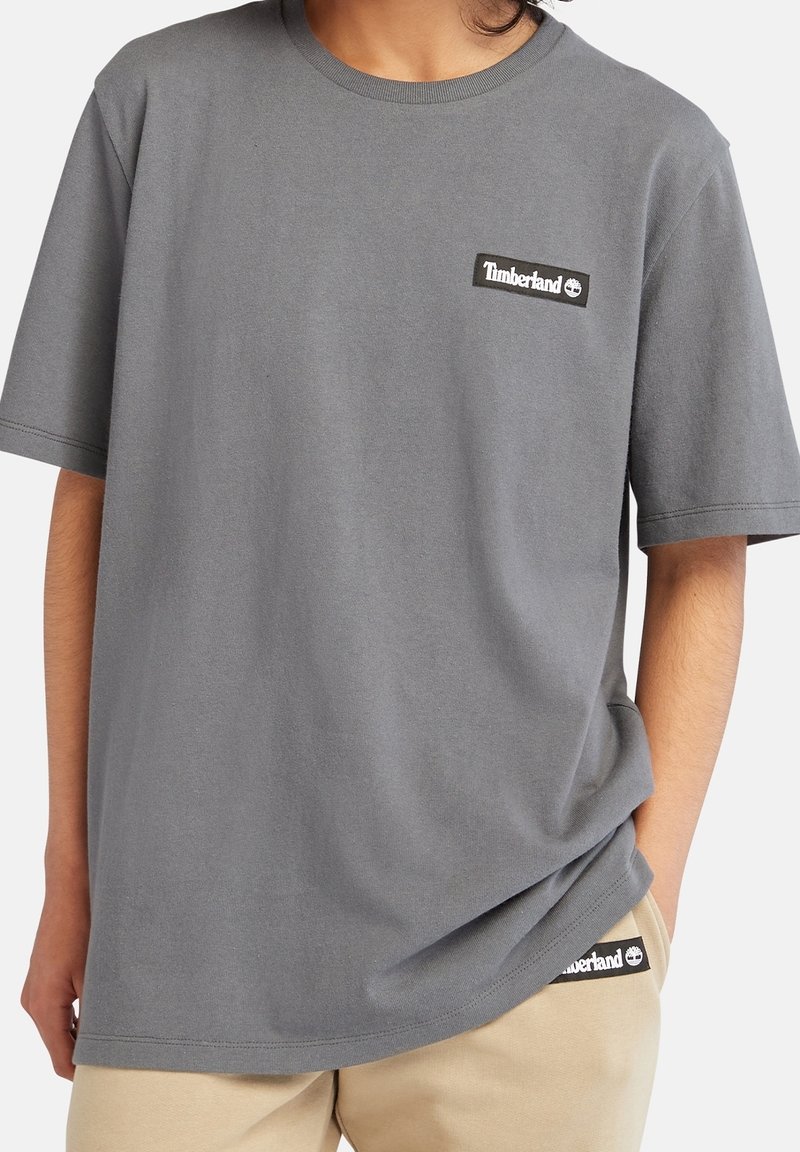 Personne portant un t-shirt Timberland gris et un pantalon Timberland beige, une main dans la poche, torse visible, fond blanc uni.