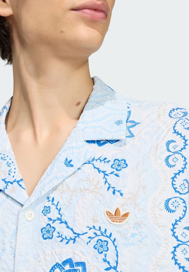 Close-up van de nek en onderkant van het gezicht van een persoon die een lichtblauw shirt draagt met blauwe bloemenpatronen en een oranje Adidas-logo op de borst.