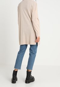 Persona vestida con un cárdigan largo beige, jeans recortados azules y botas de combate negras con cordones, de pie contra un fondo liso.