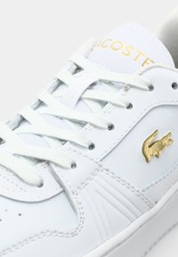 Bílá kožená teniska se zlatým detailem, s texturovaným krokodýlím logem, plochými tkaničkami a prošitými švy. Hladký povrch a polstrovaný límec.