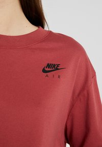 Röd bomullströja med rund halsringning. Har en svart Nike-logga och "AIR" tryckt på vänster bröstområde. Mjuk textur.