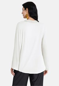 Top bianco a maniche lunghe con orlo arrotondato, realizzato in tessuto morbido. Presenta dettagli di cucitura discreti. Indossato con pantaloni neri.