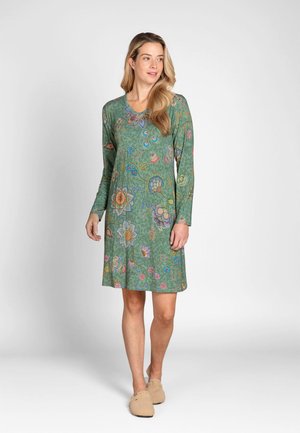 Woman wearing a green floral long-sleeve knee-length dress and beige slippers, standing against a plain light background.