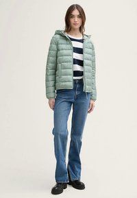 Ljust grön vadderad jacka med huva, quiltad design, dragkedja fram, kombinerad med en randig marinblå och vit topp samt utsvängda blå jeans. Svarta stövlar.