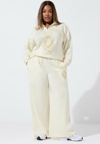 Set di felpa color crema e pantaloni a gamba larga con collo a zip e dettagli grafici dorati a forma di scudo. Texture morbida; design sportivo.