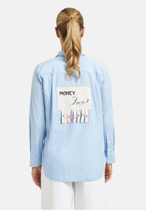 Frau mit blondem Pferdeschwanz trägt hellblaues Hemd mit Aufnäher auf dem Rücken mit der Aufschrift "MONEY OR Love?" und abreißbaren Laschen, die Dollar- und Herzsymbole zeigen.