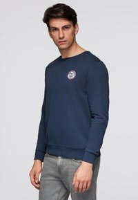 Mand med mørkt hår iført en marineblå sweatshirt med et lille rundt logo på brystet og lysegrå jeans, stående mod en ensfarvet baggrund.