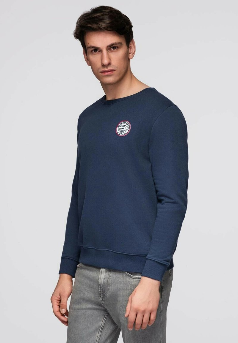Mand med mørkt hår iført en marineblå sweatshirt med et lille rundt logo på brystet og lysegrå jeans, stående mod en ensfarvet baggrund.