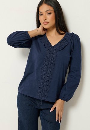Mujer con cabello largo y oscuro que lleva una blusa de manga larga azul marino con detalles de encaje y vaqueros azul oscuro, de pie contra una pared blanca.