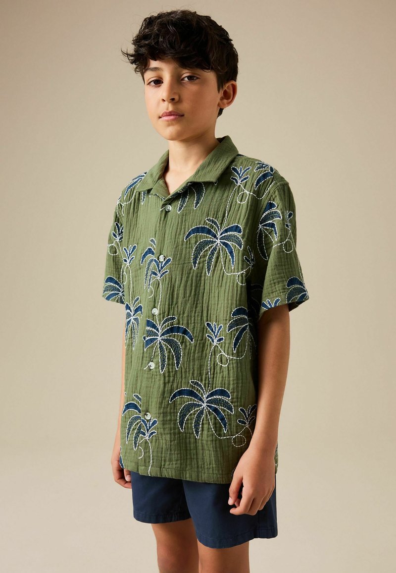 Junge in einem grünen kurzärmligen Hemd mit blauen Palmenmustern und marineblauen Shorts, stehend vor einem schlichten beigen Hintergrund.