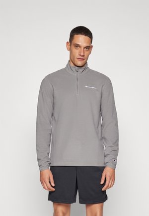 Sudadera de forro polar gris con cuello alto y cierre de una cuarta parte. Presenta un logo bordado en el lado izquierdo del pecho. Combinada con pantalones cortos negros.