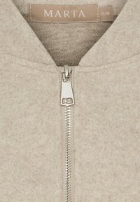 Beige jakke med lynlås lavet af blødt materiale, med en sølvfarvet lynlås, et mærke med teksten "MARTA" og størrelse S/M. Teksturen virker glat og varm.