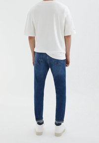 Camiseta blanca de manga corta, vaqueros ajustados azul oscuro y zapatillas blancas. Los vaqueros tienen un diseño clásico de cinco bolsillos con un ligero desgaste.