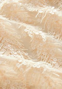 Tissu beige parsemé de sequins blancs et de franges, avec une surface texturée présentant un aspect doux et délicat.