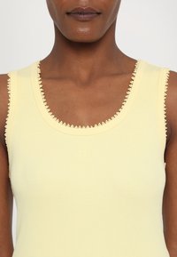 Gelbes geripptes Tanktop mit Rundhalsausschnitt, das mit einem strukturierten Spitzenbesatz an den Kanten versehen ist. Dezentes Logo auf der Brust gestickt.