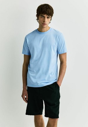 Fiatal férfi barna hajjal, világoskék rövid ujjú pólót és fekete rövidnadrágot visel, egyik kezét a zsebében tartva, egyszínű háttér előtt áll.