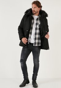 Čierna prešívaná parka s kapucňou s umelýmrúžkom, prednými zipsami, nastaviteľným šnúrkovým pásom, nosená cez károvanú flanelovú košeľu a čierne slim jeansy.