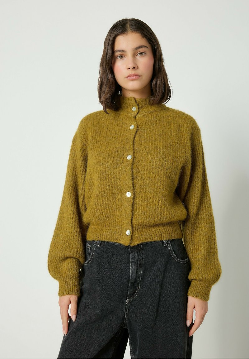 Mostardno rumeno pleteno srajco z visokim ovratnikom, sprednjim gumbom, rebrasto teksturo in napihnjenimi rokavi, v kombinaciji s črnimi kavbojkami.