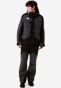 Giacca nera e grigia resistente all'acqua con cappuccio, tasche con zip e logo; abbinata a pantaloni coordinati con elementi di design angolari.
