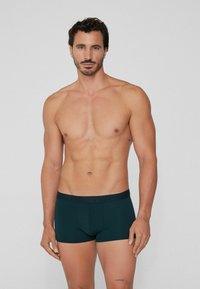 Dark green boxer shorts made of soft material with a branded elastic waistband; model shown is shirtless, standing straight.