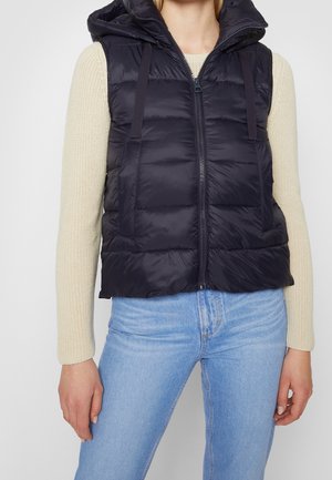 Femme portant un gilet matelassé noir à capuche par-dessus un pull beige en maille et un jean bleu clair, debout avec les bras détendus le long du corps.