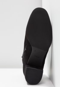 Stivale alla caviglia in suede nero con suola in gomma liscia, tacco a blocco e zip laterale, fotografato dal lato della suola su uno sfondo bianco.