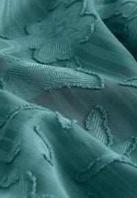 Tessuto turchese ricamato con motivi floreali, caratterizzato da accenti texture e aree trasparenti, crea un aspetto morbido e stratificato.
