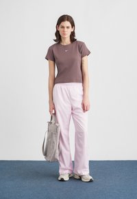 Νεαρή γυναίκα στέκεται σε μπλε χαλί, φορώντας καφέ μπλουζάκι, ανοιχτό ροζ παντελόνι, λευκά αθλητικά παπούτσια, κρατώντας γκρι τσάντα κατά μήκος λευκού τοίχου.