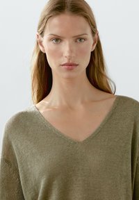 Olivgrüner V-Ausschnitt-Pullover mit einer weichen Strickoberfläche, der eine lässige Passform und subtile Nähte an Hals- und Ärmelansatz aufweist.