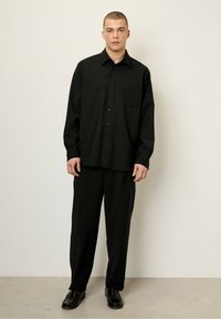 Chemise noire à manches longues avec fermeture à boutons, poche sur la poitrine, associée à un pantalon noir. Texture lisse, coupe décontractée, design simple. Chaussures noires.