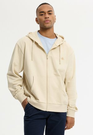 Hombre con sudadera con capucha beige con cremallera encima de una camisa azul clara y pantalones oscuros, de pie con una mano en el bolsillo sobre un fondo liso.