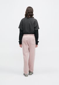 Άτομο με μαλλιά μέχρι τους ώμους στέκεται γυρισμένο προς τα πίσω, φορώντας ένα μαύρο πολυεπίπεδο μπλουζάκι, ανοιχτό ροζ φόρμες και γκρι αθλητικά παπούτσια σε λευκό φόντο.