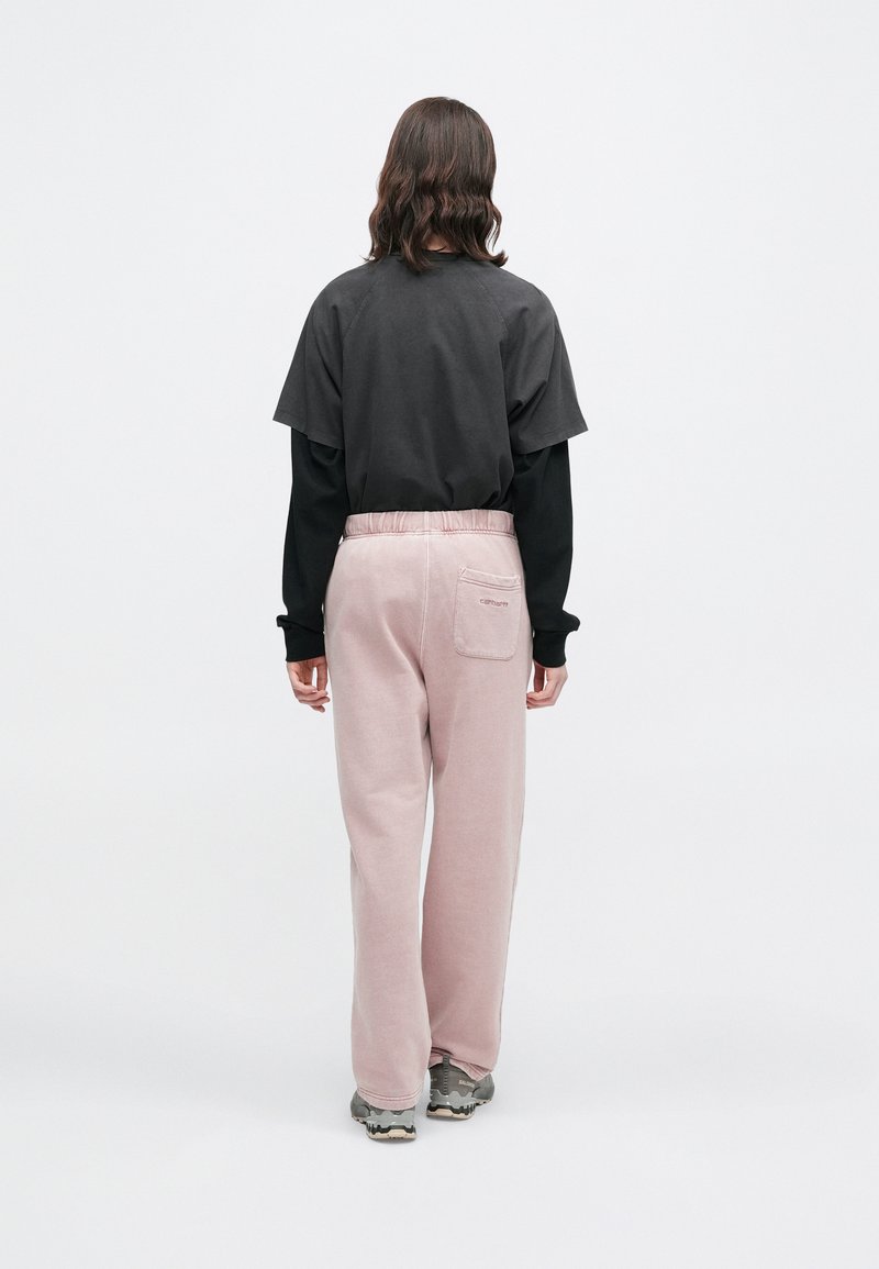 Persona con capelli di lunghezza media che sta voltata, indossando un top nero a strati, pantaloni della tuta rosa chiaro e scarpe da ginnastica grigie su uno sfondo bianco.