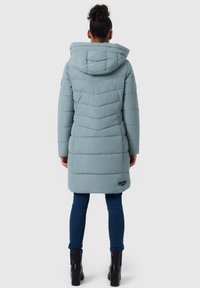 Gewatteerde middellange jas in lichtblauw met een capuchon, voorzien van een chevronsteek en een logo-opdruk nabij de zoom. Draagt een donkere jeans en laarzen.