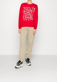 Felpa rossa con design di fiocchi di neve bianchi e testo "Baby it's cold Outside", abbinata a pantaloni jogger beige e sneakers bianco-neri.