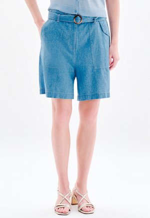 Femme portant un short bleu ceinturé avec poches avant et un haut bleu clair, debout en sandales à talons beiges sur fond blanc.