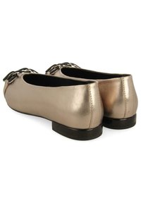Chaussures plates métalliques dorées avec un bout rond et un petit nœud noir, présentant une texture lisse et un petit talon noir.