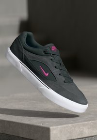 Tamsiai pilkas Nike sportbaigis su rožiniu logotipu ir baltu padu, levituojantis virš pilkos akmens dangos, prieš neryškų foną.