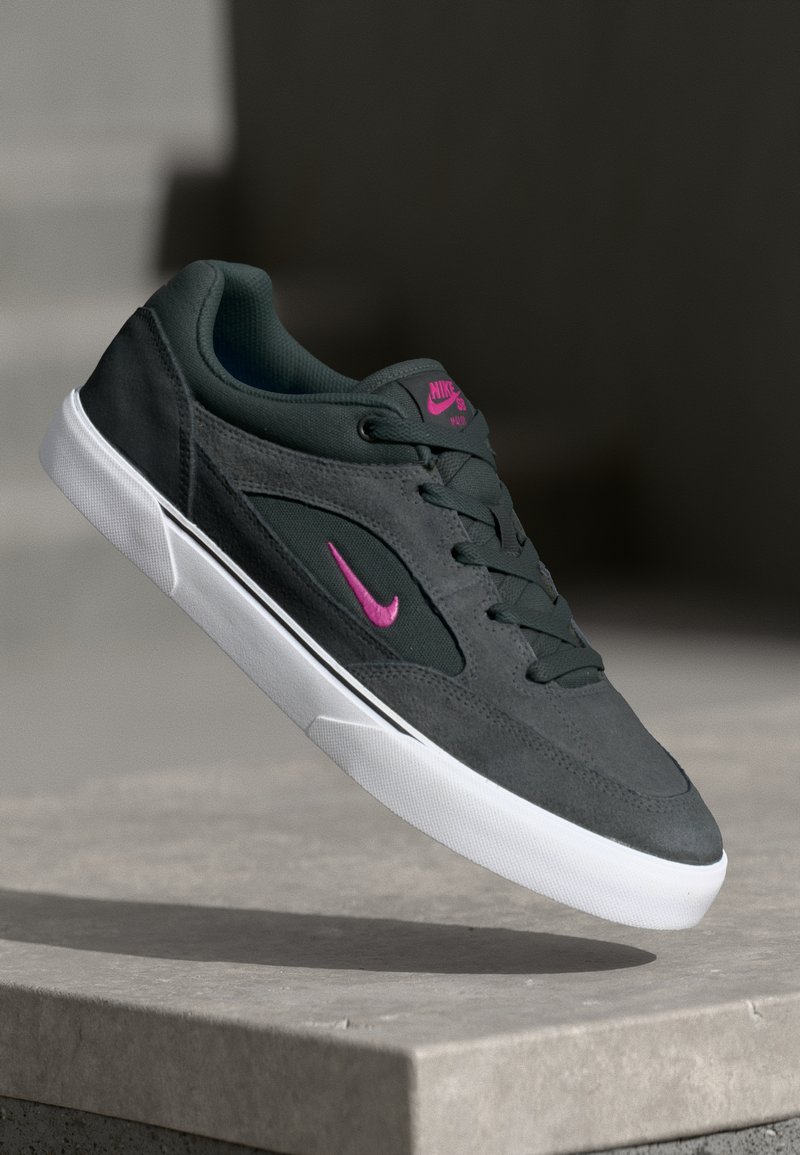 Tamsiai pilkas Nike sportbaigis su rožiniu logotipu ir baltu padu, levituojantis virš pilkos akmens dangos, prieš neryškų foną.