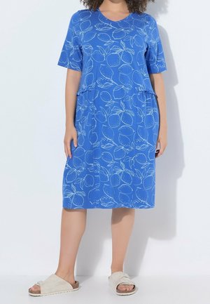 Mujer con un vestido azul hasta la rodilla con estampados blancos de limón y sandalias beige abiertas en los dedos, de pie sobre un fondo blanco.