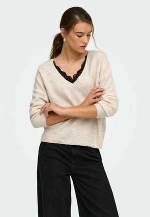 Donna che indossa un maglione beige a scollo a V sopra una maglia nera in pizzo e pantaloni neri, guarda verso il basso con le braccia incrociate su sfondo uniforme.