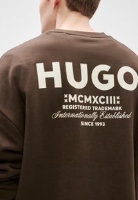 Felpa marrone con grande scritta bianca "HUGO" e ulteriori dettagli di branding sotto. Il testo è in grassetto con una texture leggermente in rilievo.