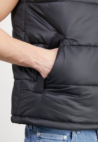 Gilet nero imbottito con sezioni trapuntate, tasca laterale e una superficie liscia. Realizzato in materiale leggero e isolante.