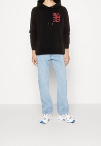 Felpa nera con stampa grafica rossa, tasca a marsupio, abbinata a jeans dritti azzurri e sneakers bianche con accenti blu.