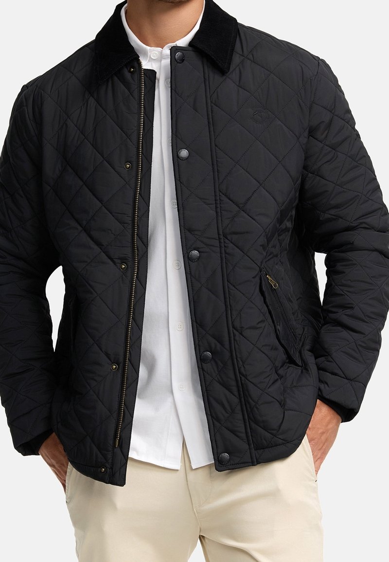 Homme portant une veste matelassée noire avec des boutons-pression sur une chemise blanche à boutons et un pantalon beige, les mains dans les poches de la veste.
